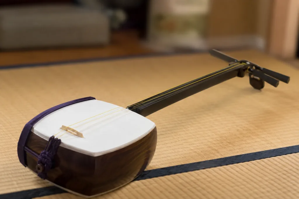 A shamisen instrument with a long neck and a rectangular body, played with a plectrum in traditional Japanese music style.